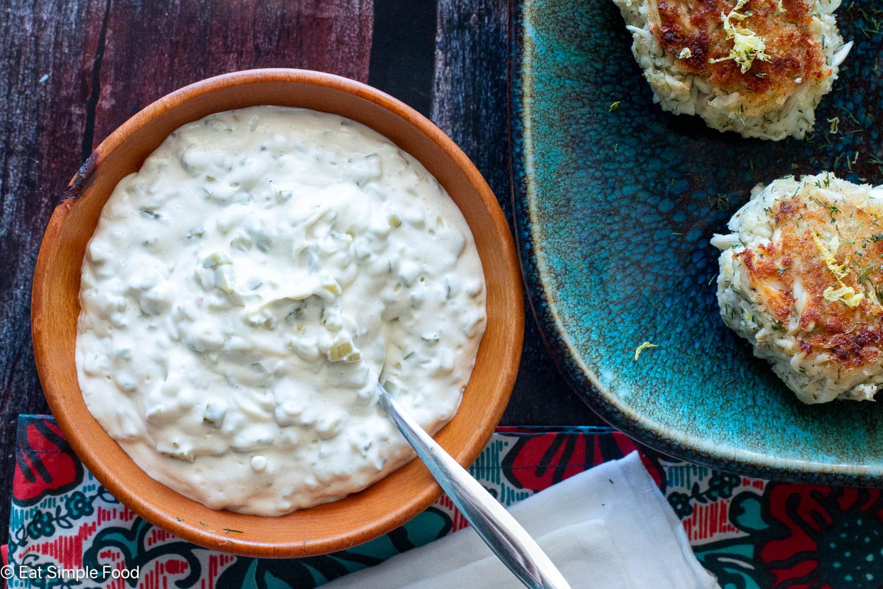 Top view of wood bowl filled with white mayonnaise sauce with pickles. Crab cakes on aqua plate on the side.