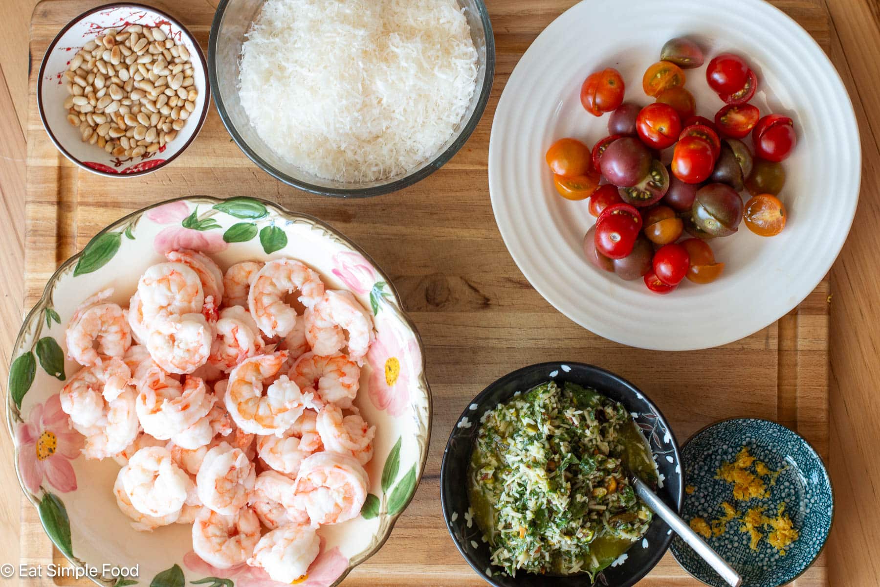 Top down view of a wood cutting board with a bowl of poached shrimp, a plate of halved cherry tomatoes, basil pesto, pine nuts, and grated Parmesan cheese.