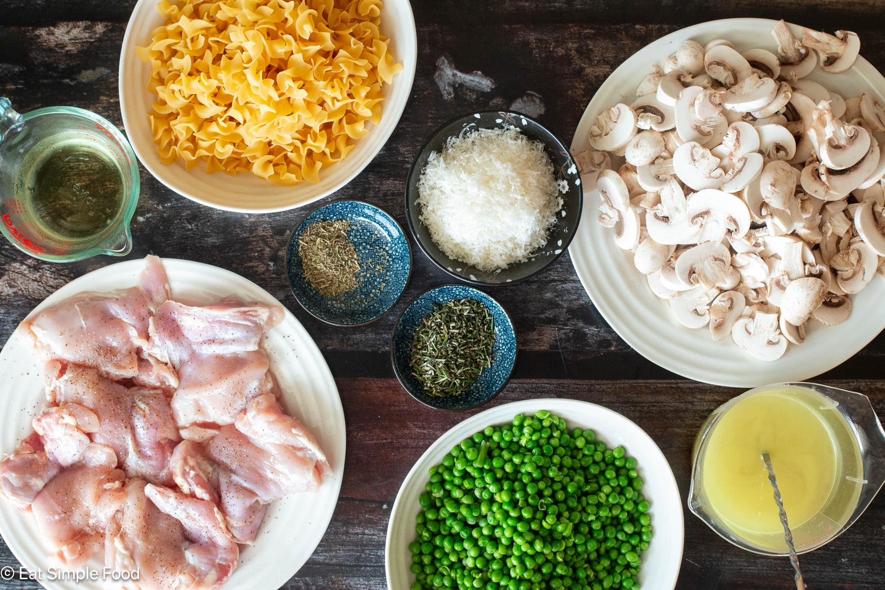 Ingredients on a wood table: bowl of green peas, plate with raw chicken thighs, plate of sliced mushrooms, bowl of uncooked egg noodles, chicken broth cup, ramekins of herbs and cheese.