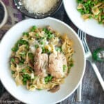 Top down view of a white plate with chicken over egg noodles and peas. Forks and parmesan cheese on side.