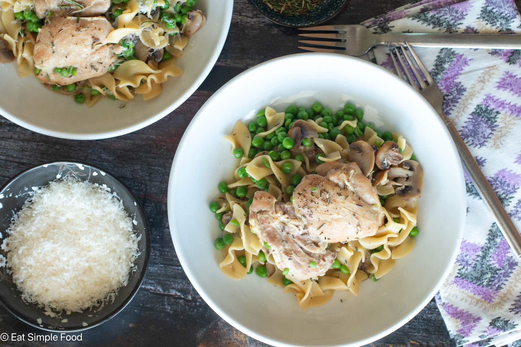 Top down view of one plate with chicken, mushrooms, peas, and egg noodles. Side of Parmesan cheese on the side. Forks and purple napkin on side.