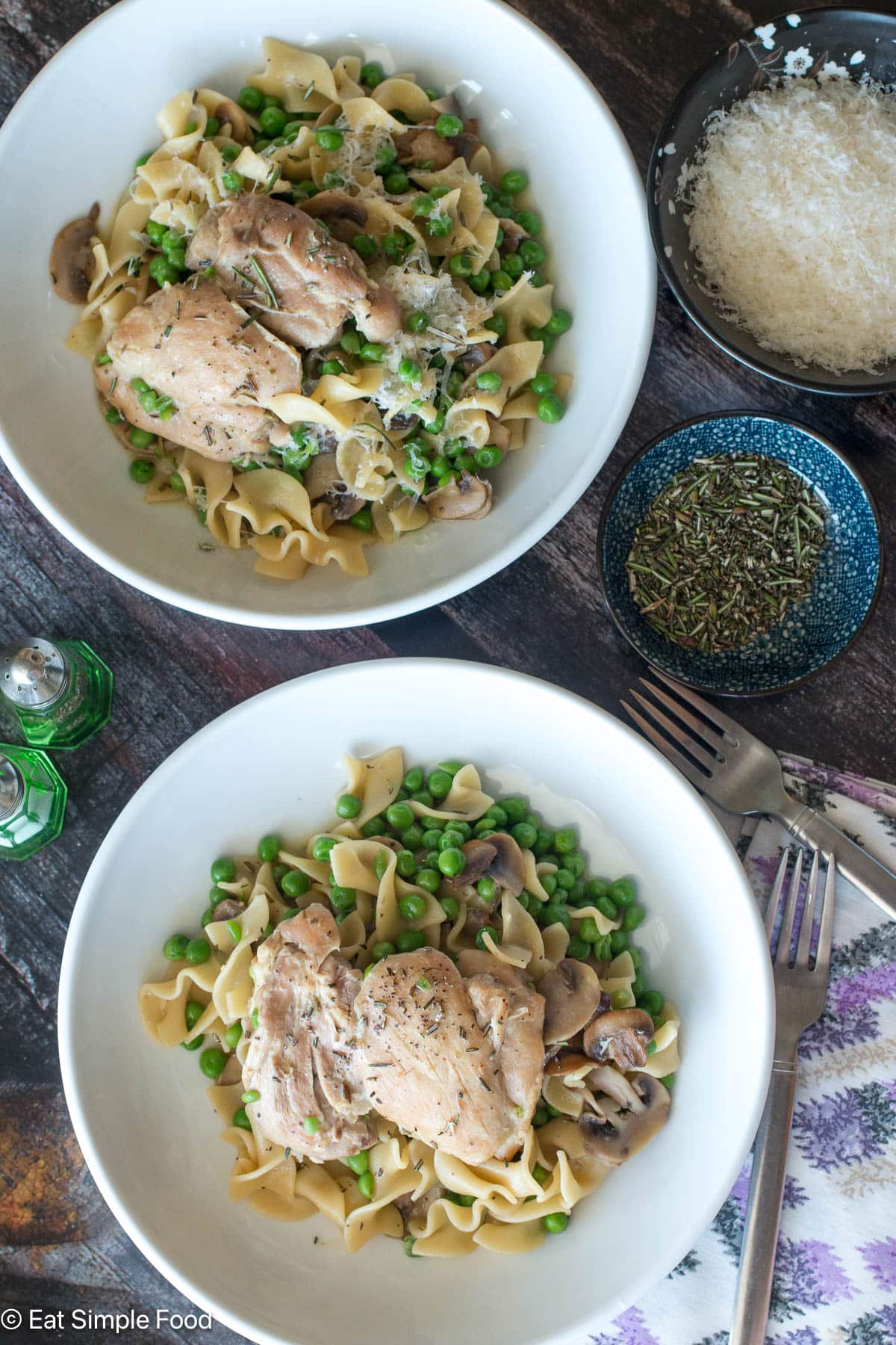 Top down view of two plates with chicken, mushrooms, peas, and egg noodles. Side of rosemary and Parmesan cheese on the side.
