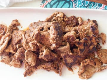 White plate with browned meat slices with colorful napkin in background.