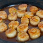 Side view of brown plate of cooked scallops in an orange brown sauce with zest on top.