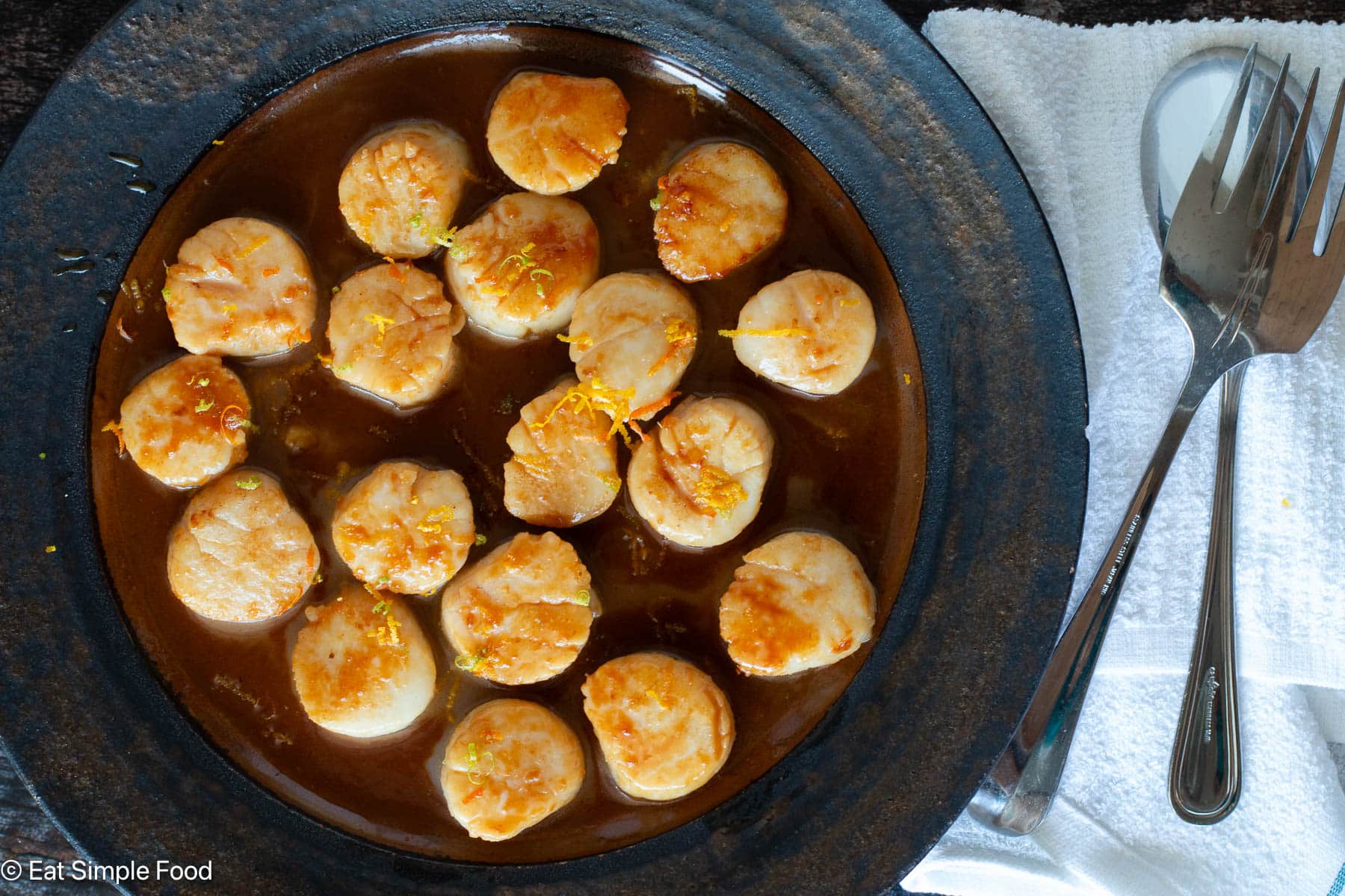Top down view of cooked scallops on a brown plate with fork and spoon on a white cloth.