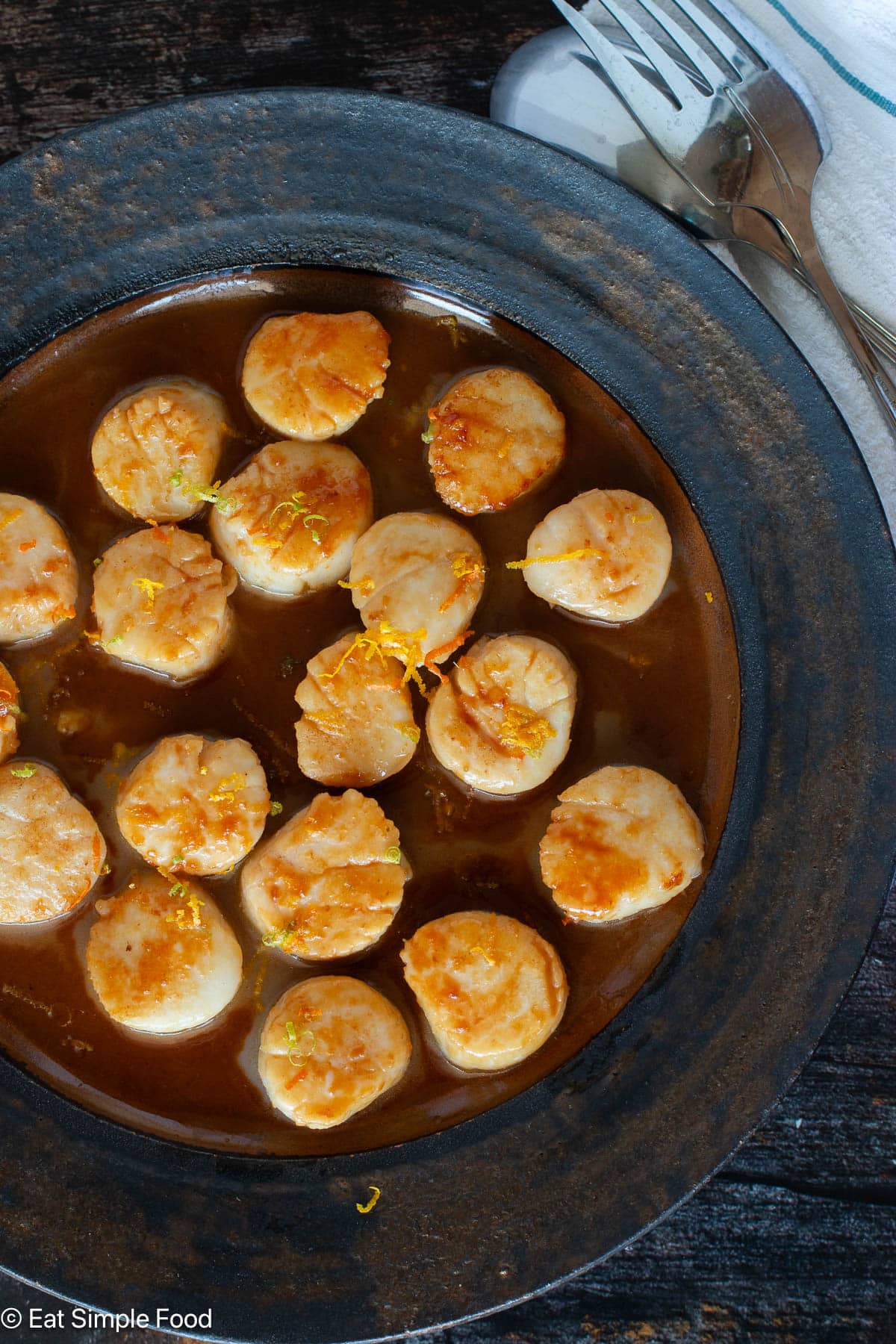 Top down view of cooked scallops on a brown plate with silverware in background.