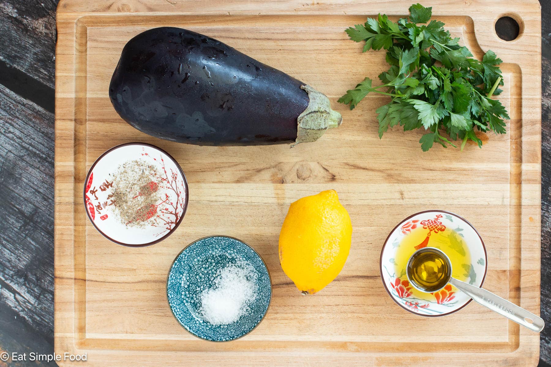 Ingredients on a wood cutting board: raw eggplant, lemon, parsley sprigs, salt, pepper, and olive oil with a Tablespoon.