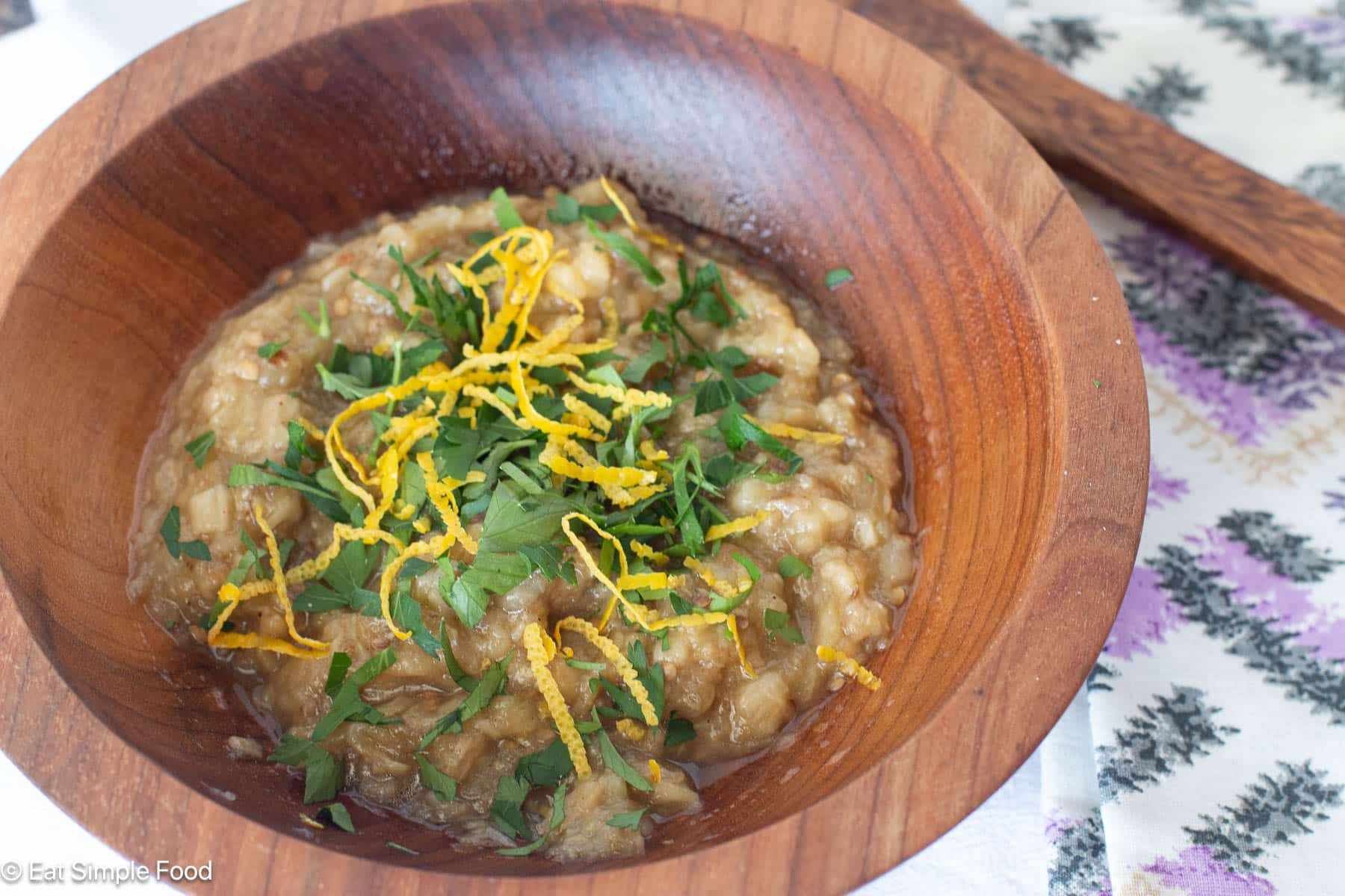 Wood bowl of brown dip garnished with chopped parsley and lemon zest. Purplee and white napkin in background. Side view.
