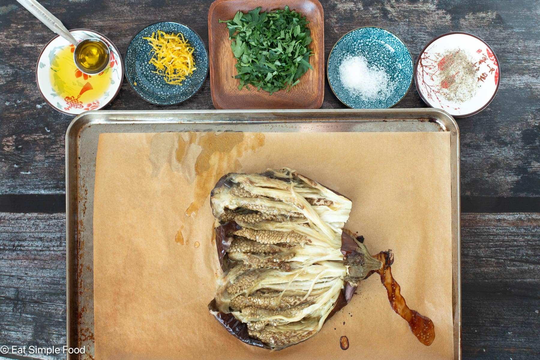 Cooked Eggplant cut in half on a parchment lined sheet pan with different bowls of ingredients in the background. top down view.