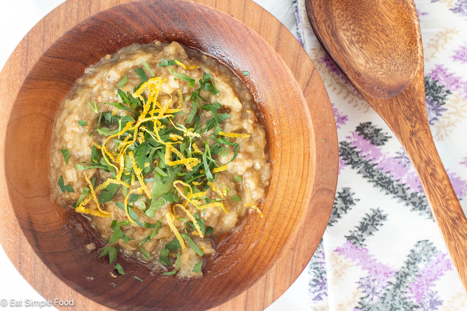 Wood bowl of brown dip garnished with chopped parsley and lemon zest. Purplee and white napkin in background. Top view.