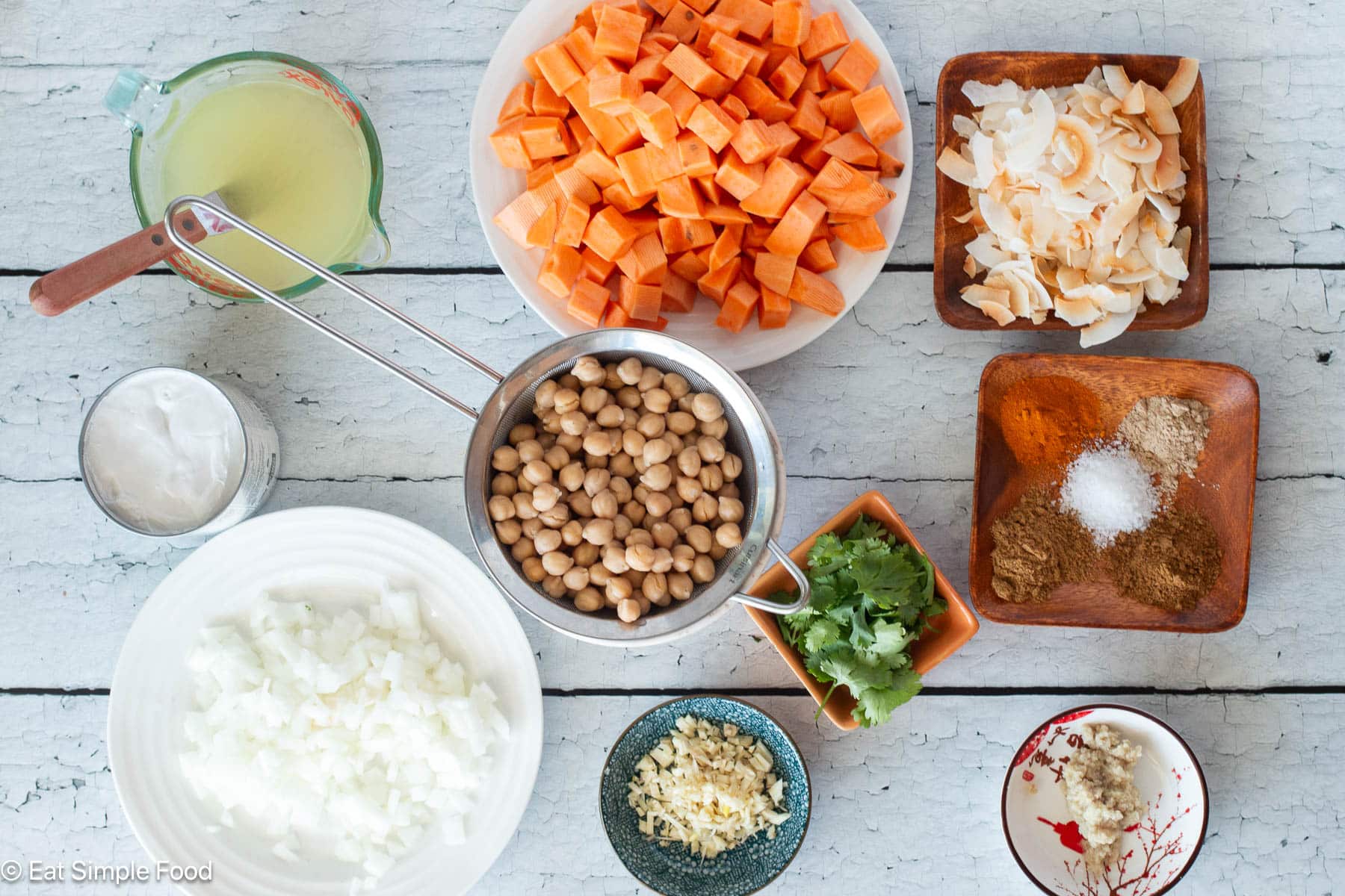 Top down view of ingredients on a white table: diced onions, chickpeas, cilantro, garlic, ginger, colorful spices, coconut flakes, and diced sweet potatoes.