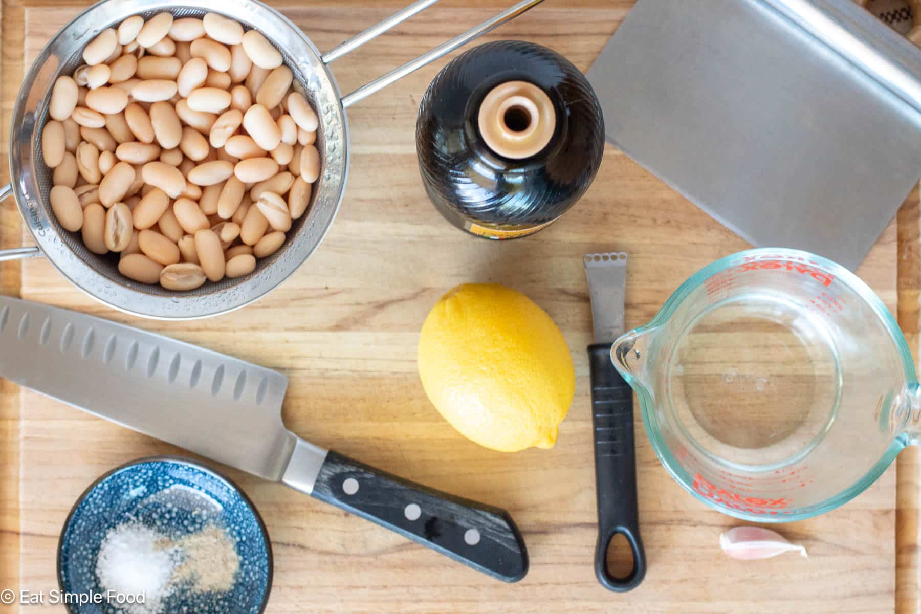 Ingredients on a wood cutting board with a chefs knife, pastry scraper, and lemon zester. Drained white beans, salt and pepper, a garlic clove, and a whole lemon.