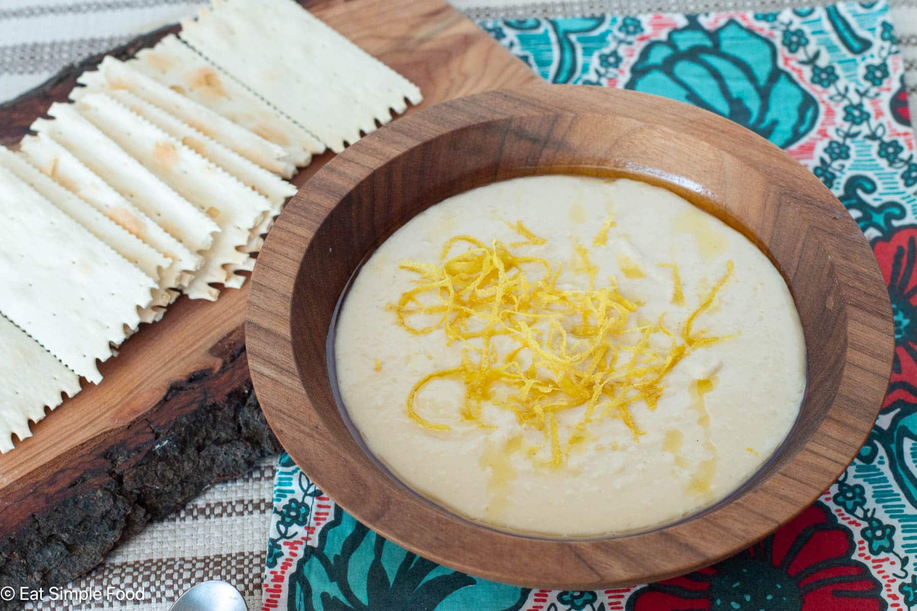 Side view of pureed white dip with lemon zest on wood bowl. Crackers in background.