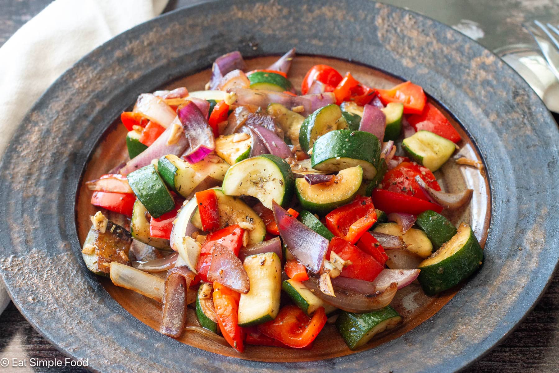 Side view of chunky cooked onions, zucchini, peppers on a brown plate with white napkin in background.