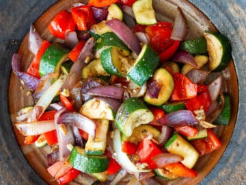 Top down view of chunky cooked onions, zucchini, peppers on a brown plate with white napkin in background. Close up.