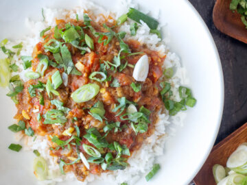 Top down view of white plate with rice and red curry garnished with sliced green onions and green herbs. Silverware on the side. Close up.