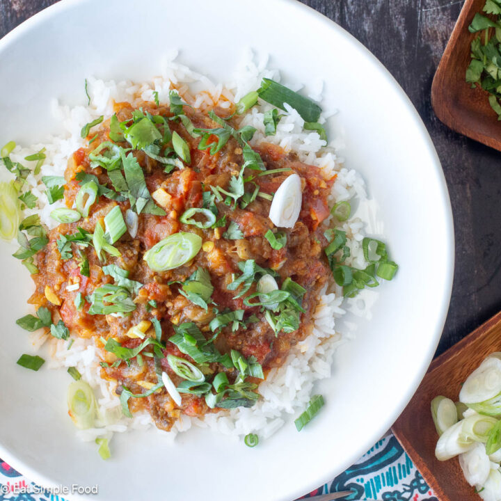 Top down view of white plate with rice and red curry garnished with sliced green onions and green herbs. Silverware on the side. Close up.