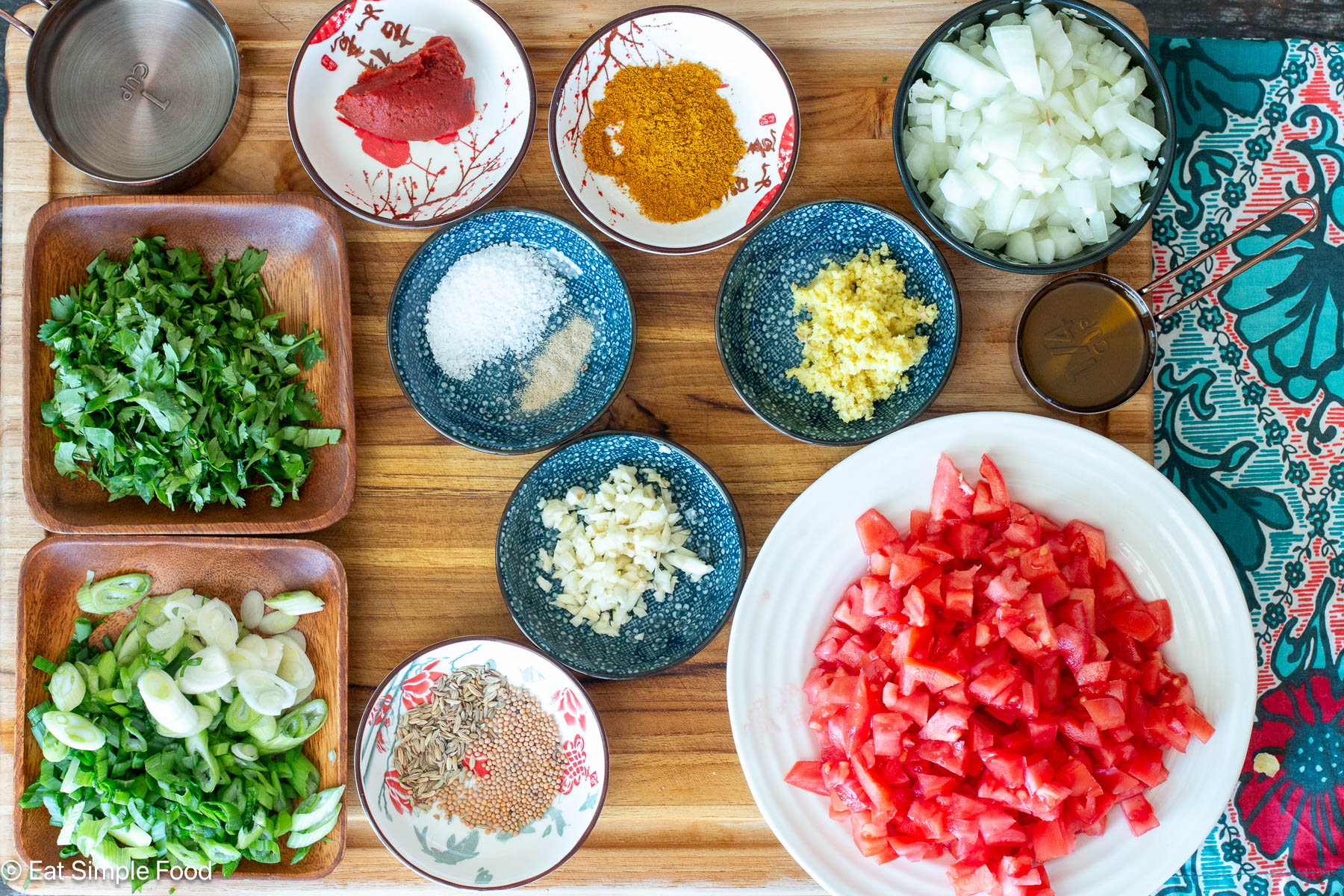 Ingredients on a wood cutting board: cilantro, sliced onions, diced tomatoes, diced onions, ginger, garlic, tomato paste, and saucers of spices.
