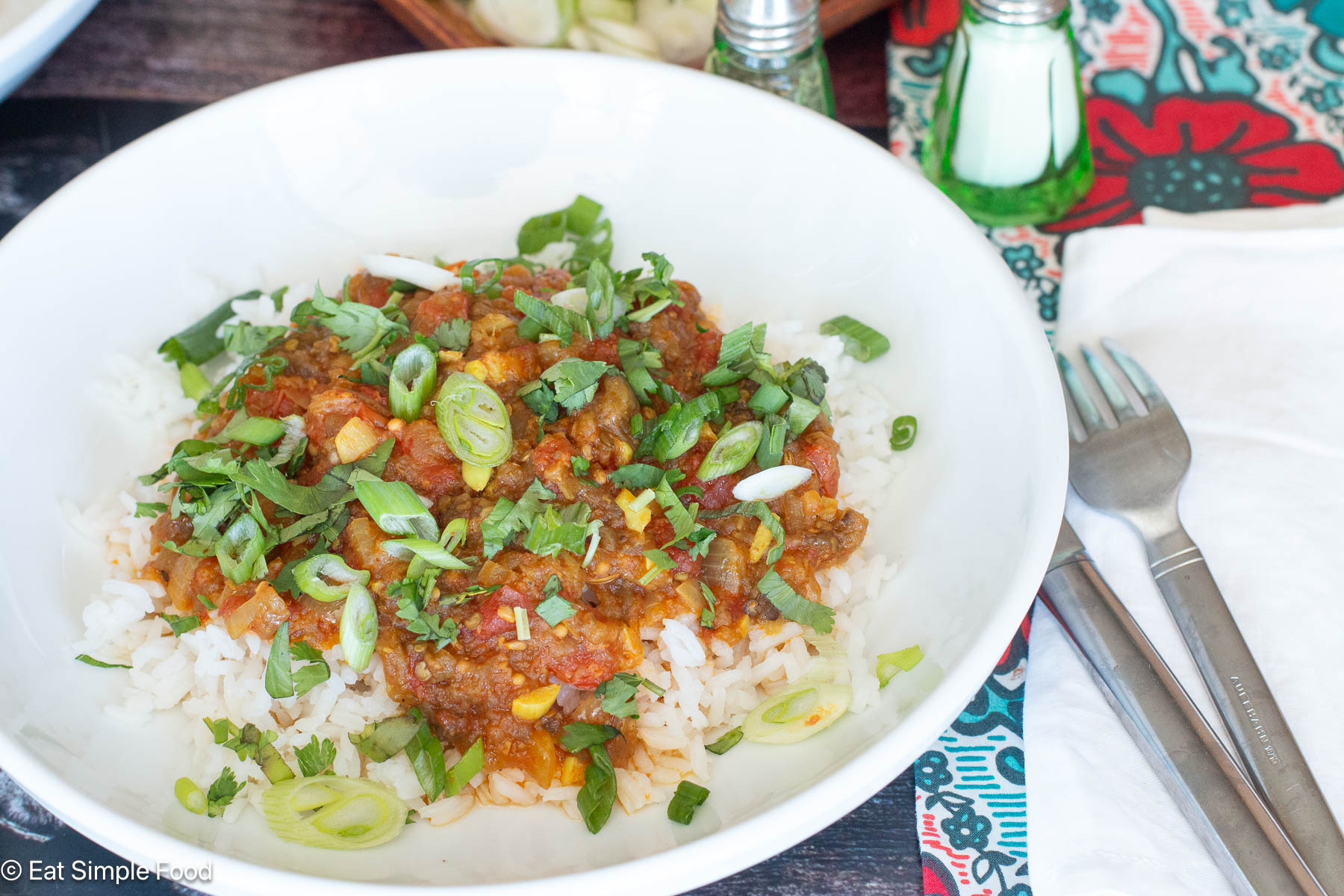 Red sauce over rice topped with sliced green onions and herbs on a white plate. Silverware on the side.