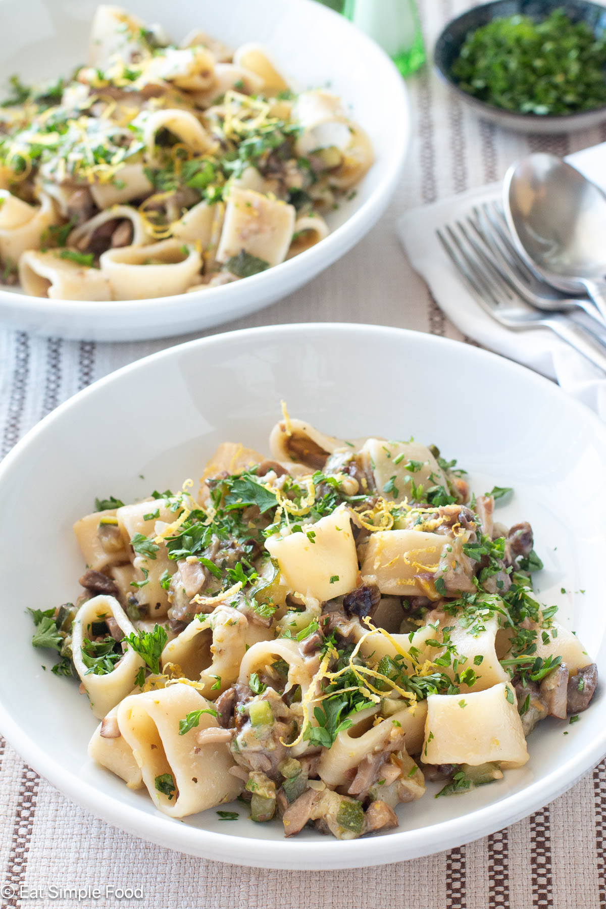 Two white plates filled with pasta and ingredients with herbs on top. Silverware on side.