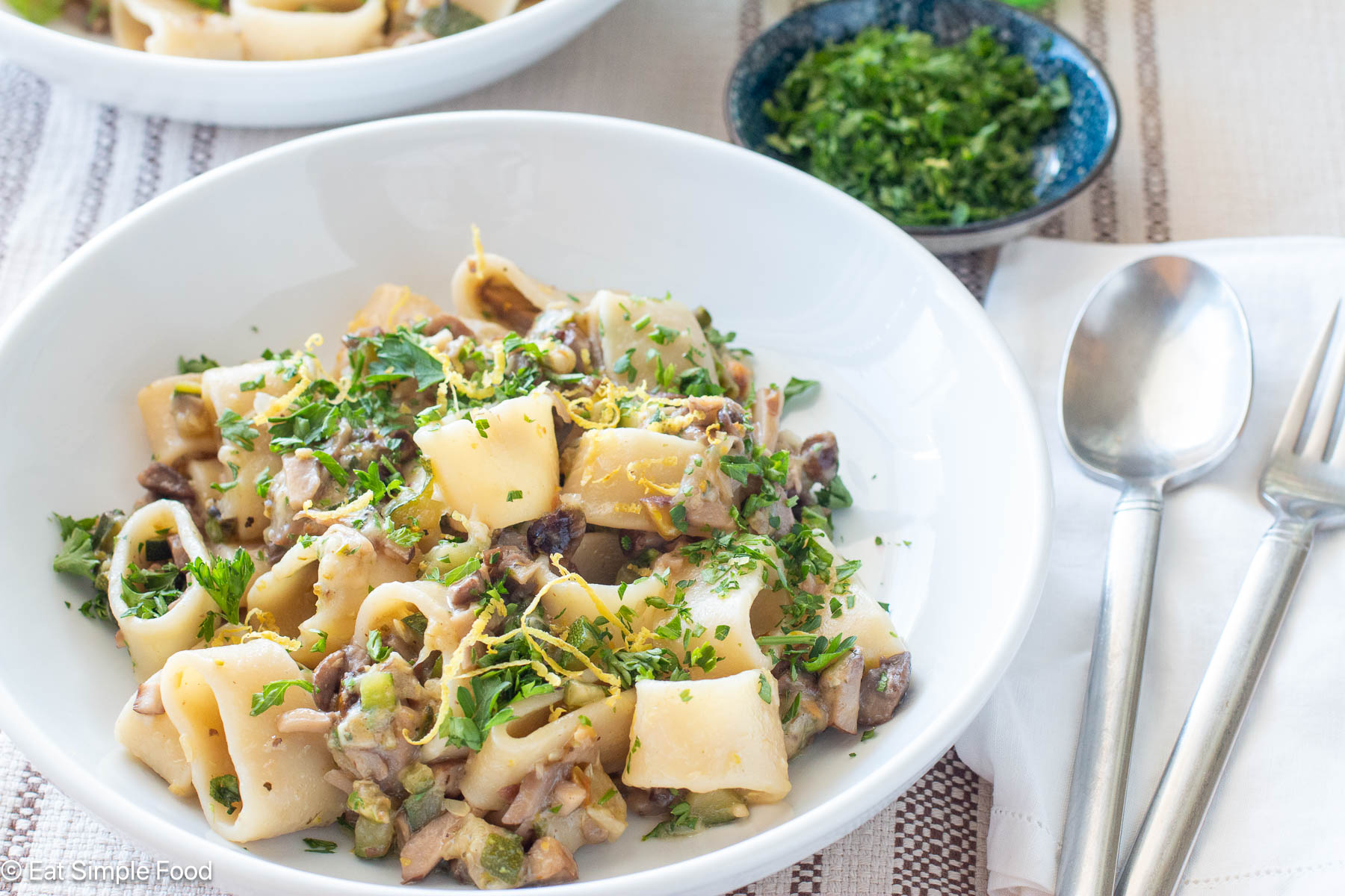 Side view of pasta on a white plate topped with lemon zest and green parsley in the background. Silverware on the side.