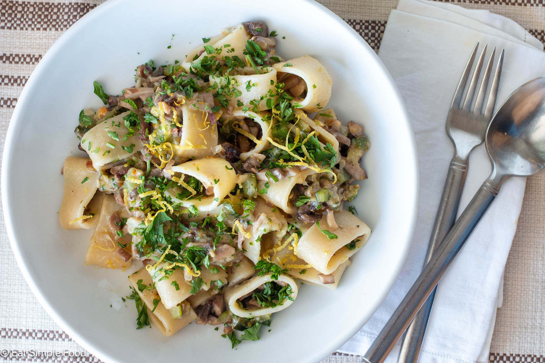 Top down view of pasta on a white plate topped with lemon zest and green parsley in the background. Silverware on the side.