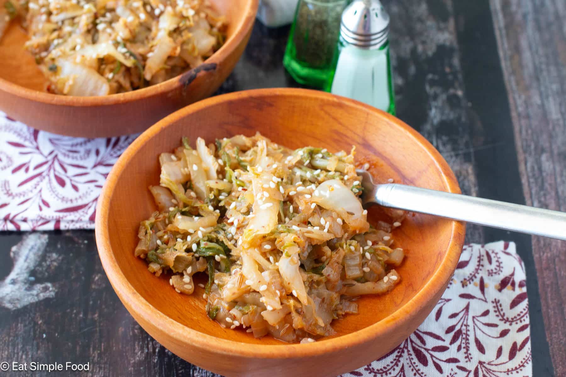 Close up of wood bowl filled with cooked cabbage slices and topped with sesame seeds with a fork in the bowl. Napkins on the side.