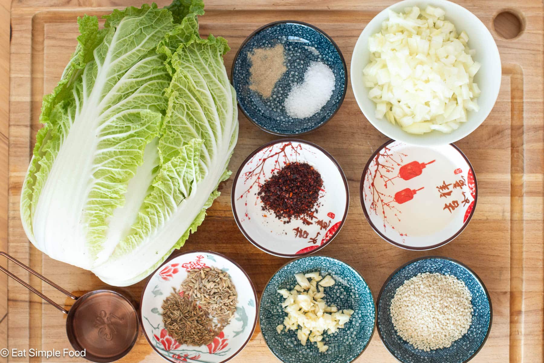 Ingredients on a wood cutting board: whole napa cabbage, containers containing diced onions, minced garic, herbs and spices.