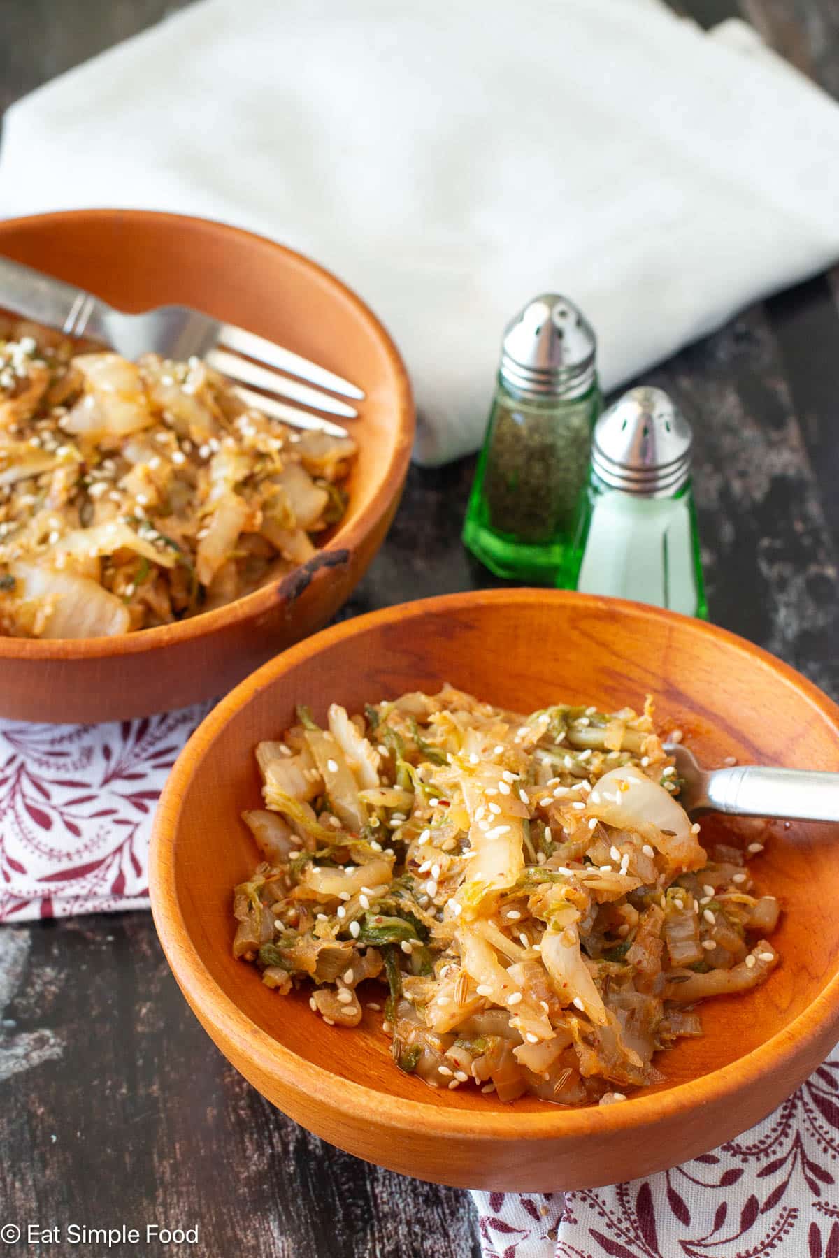 Two wood bowls filled with cooked cabbage topped with sesame seeds. Napkins and salt and pepper shakers in the background.