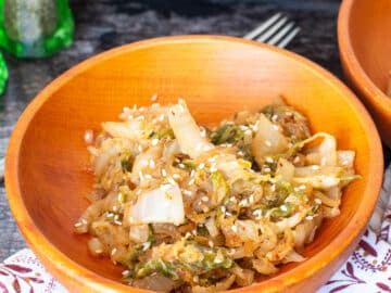Close up of wood bowl filled with cooked cabbage slices and topped with sesame seeds with a fork in the bowl. Salt and pepper shakers in background.