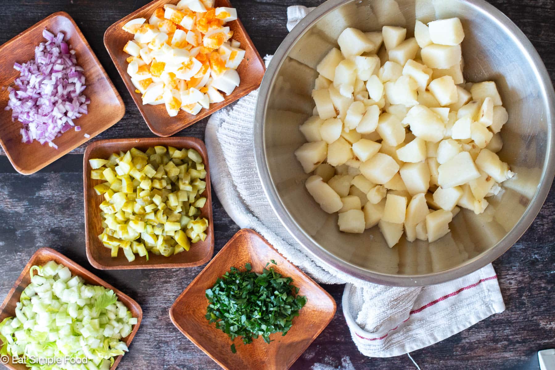 Ingredients on a wood table: bowl of chunked peeled potatoes, wood plates of diced red onions, herbs, diced pickles, diced celery, and diced boiled eggs.