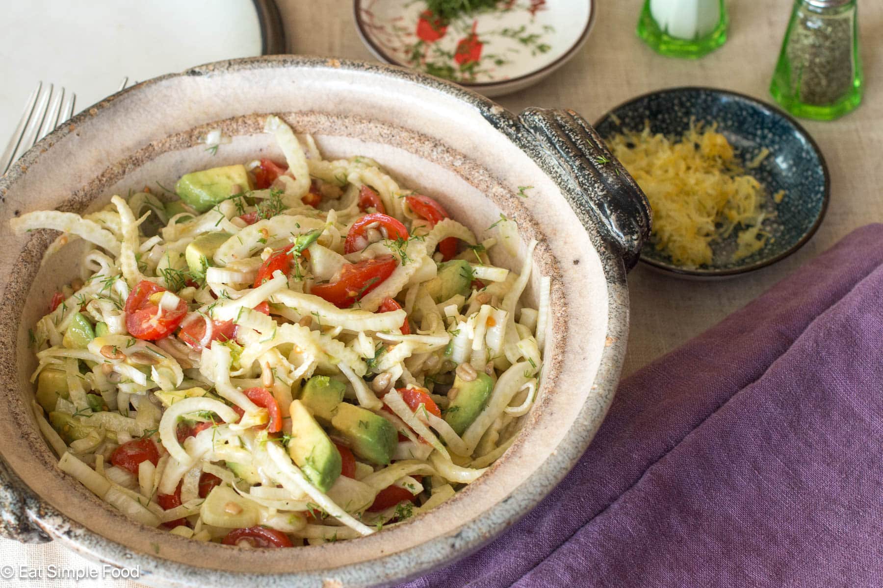 Brown bowl of fennel salad with tomatoes and avocado with purple napkins on the side.
