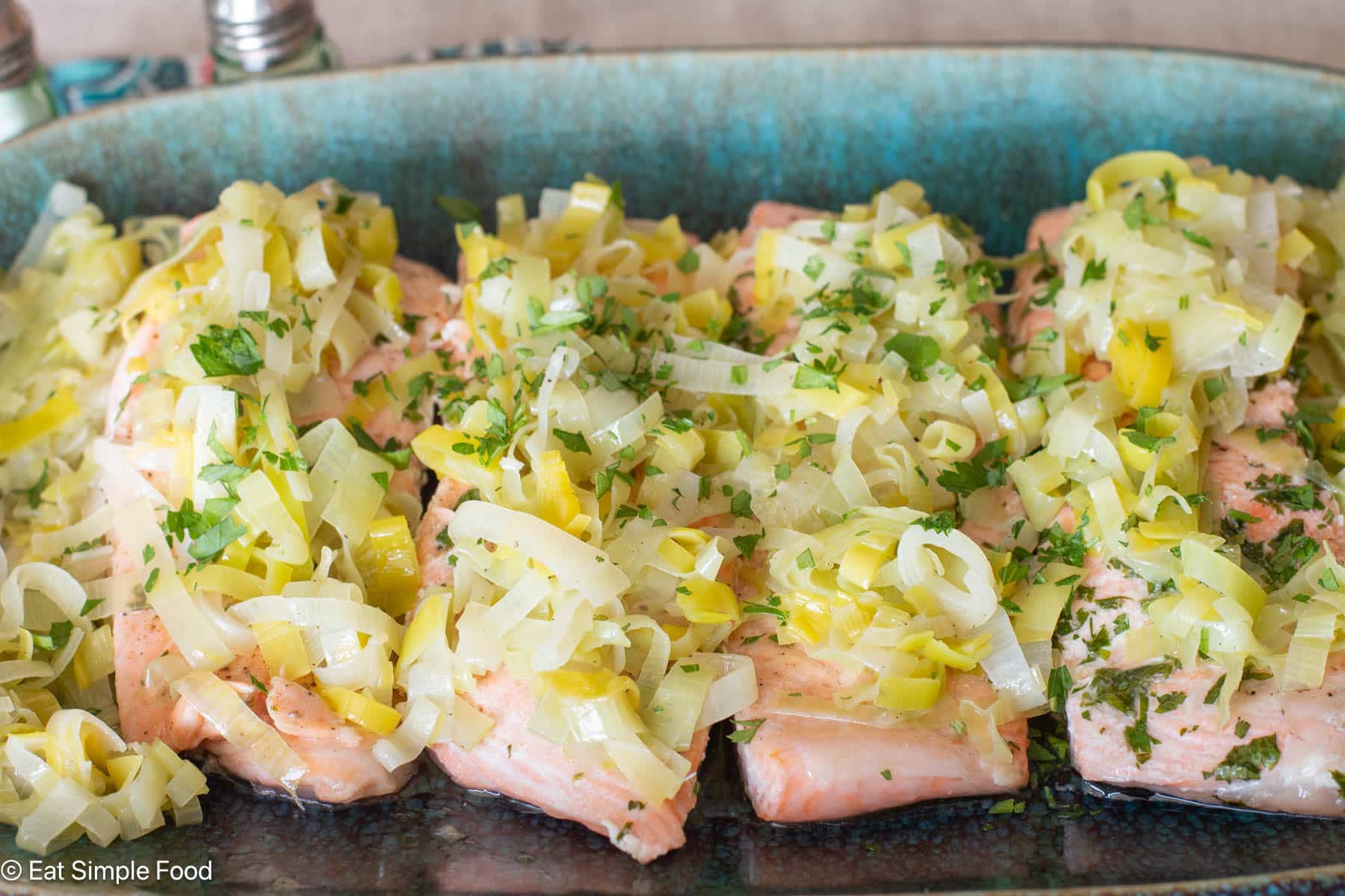 4 pieces of salmon on an aqua platter with sliced cooked leeks on top. Side view.