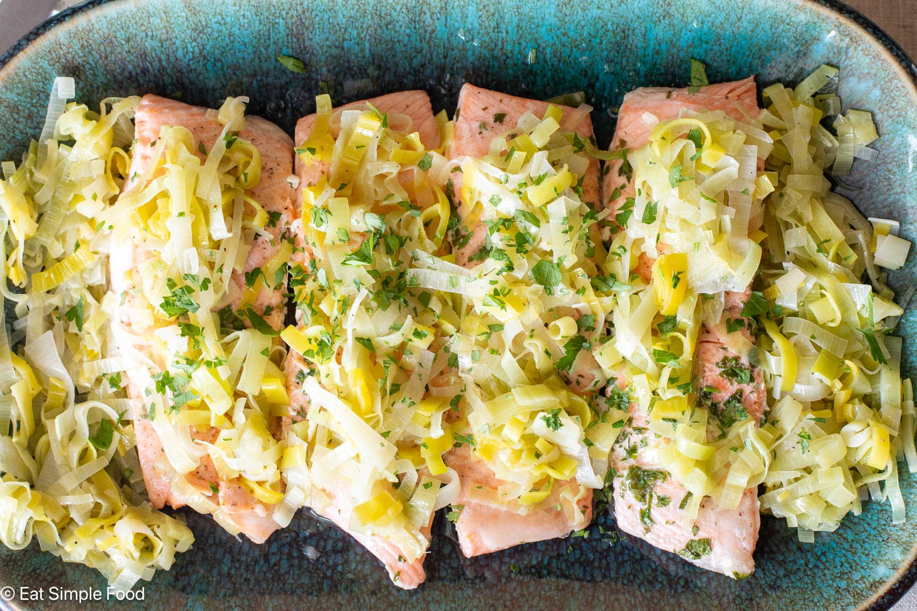 4 pieces of salmon on an aqua platter with sliced cooked leeks on top. Top view.