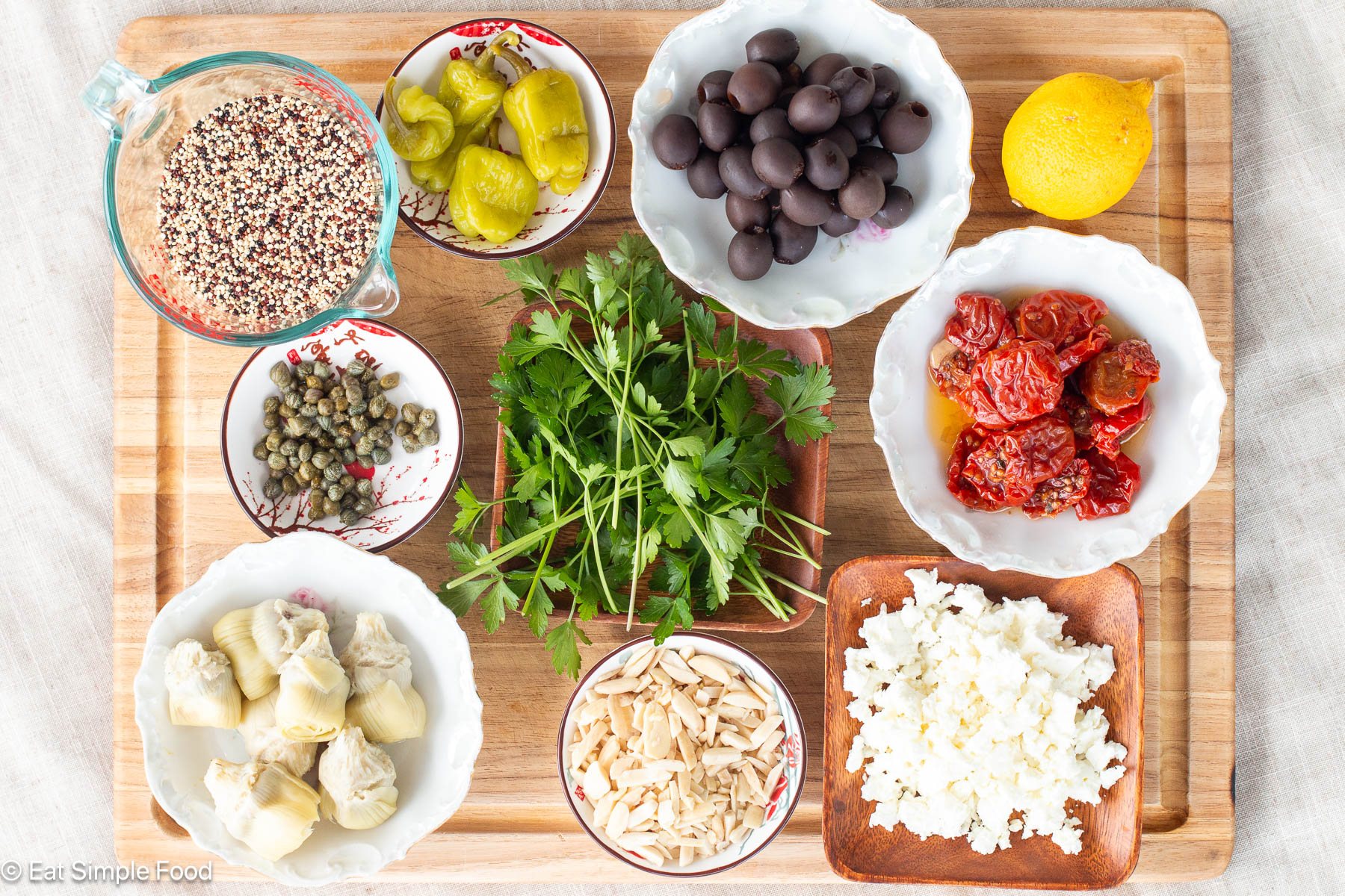 Top down view of ingredients on a wood cutting board: olives, sun dried tomatoes, feta, artichokes, capers, quinoa, peppers, parsley, almonds, and lemon.