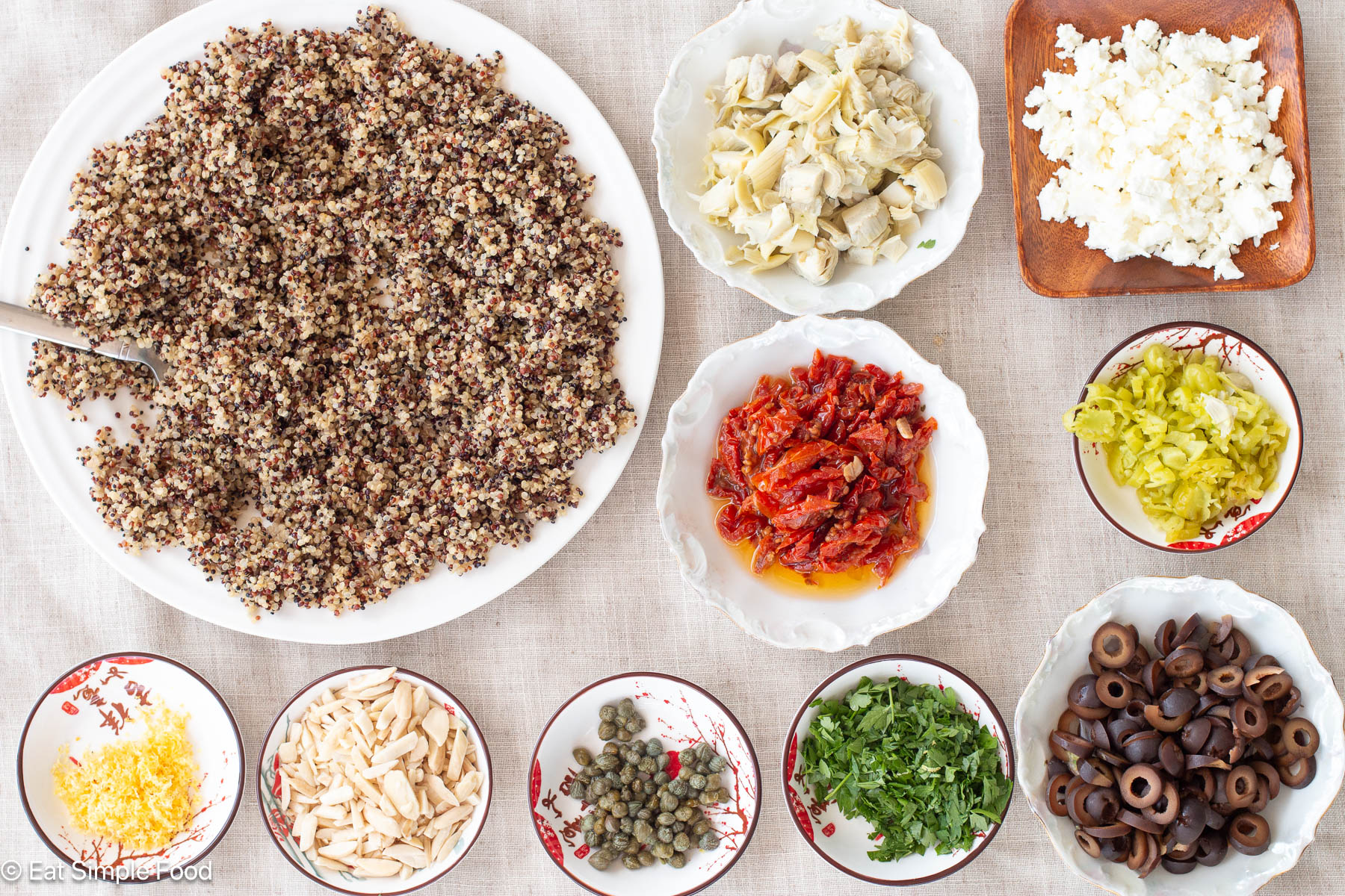 Top down view of chopped up ingredients on a wood cutting board: olives, sun dried tomatoes, feta, artichokes, capers, quinoa, peppers, parsley, almonds, and lemon.