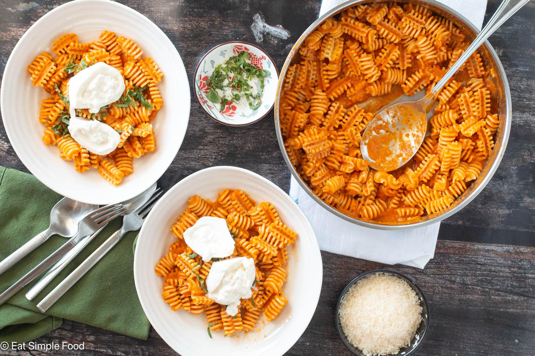 Top down view of pasta in red sauce with two plates of pasta with cheese on top, green napkins and silverware on the side.