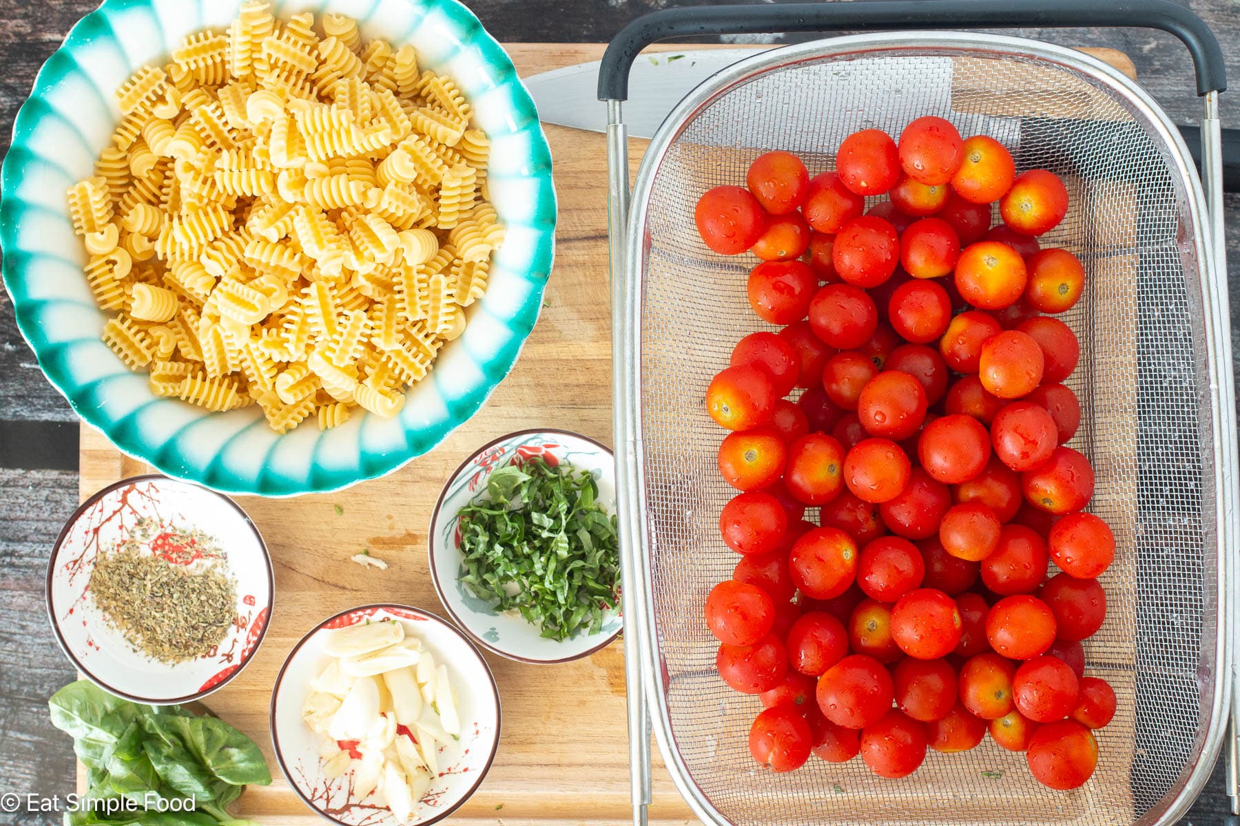 Top down view of ingredients on a wood cutting board: cherry tomatoes in strainer, sliced basil, garlic cloves, seasoning, and dry pasta.