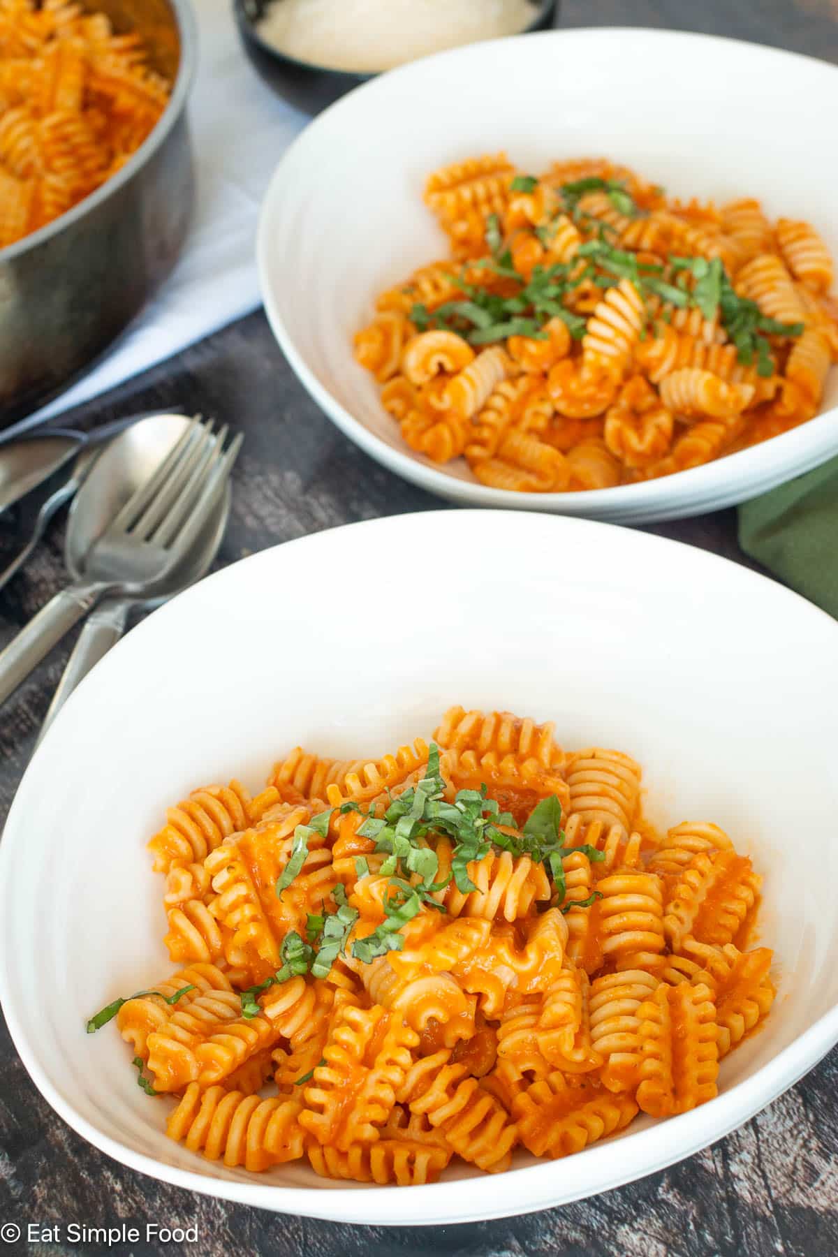 Side view of 2 plates of pasta in tomato sauce in a white plate with basil on top and a green napkin and silverware.