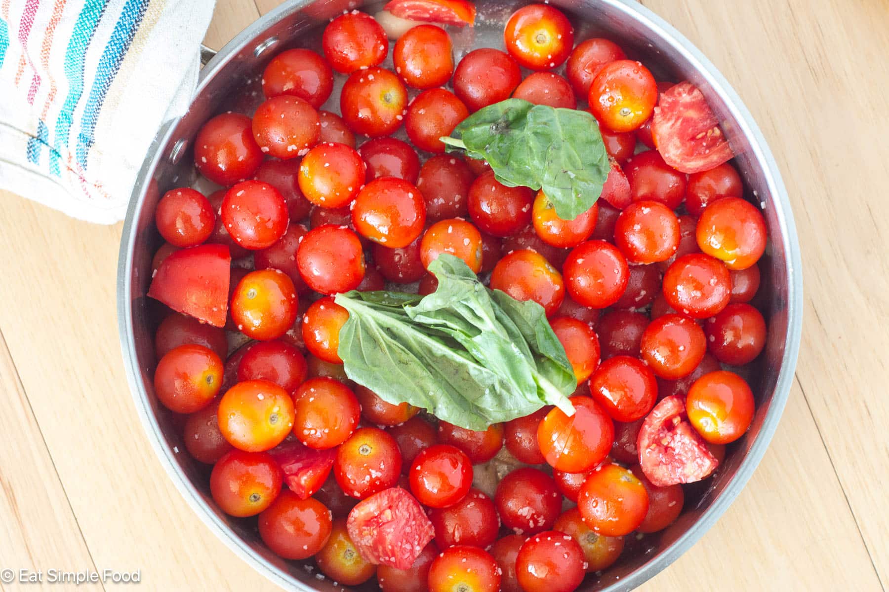 Whole cherry tomatoes and basil leaves with salt in a stainless steel pan.